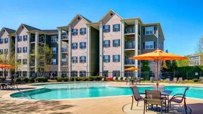 A wide shot of the expansive, curving community swimming pool on a sunny day. The clubhouse and apartment buildings are visible in the background.