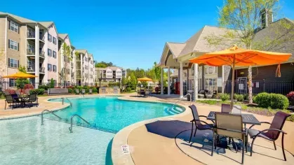 A three-story apartment building with balconies, viewed from the community pool deck. A patio table and orange umbrella are in the foreground.