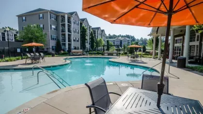 A seating area with an orange umbrella on the pool deck. The curving swimming pool and community clubhouse are visible on a clear day.