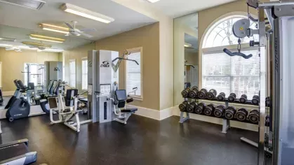 The community fitness center, featuring a dumbbell rack, Cybex weight machines, and cardio equipment. A large mirror and arched window are in the background.