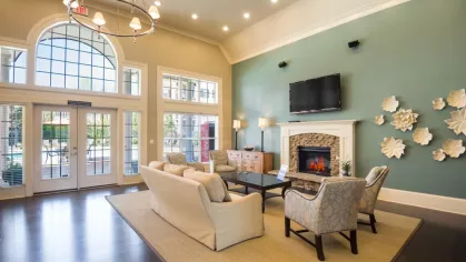 The resident lounge in the community clubhouse, featuring a comfortable seating area with a sofa and armchairs. A fireplace and wall-mounted TV are set against a teal accent wall.