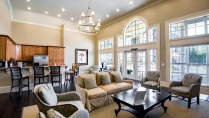 The spacious community clubhouse features a resident lounge with sofas and armchairs. A full kitchen with a breakfast bar is visible in the background.