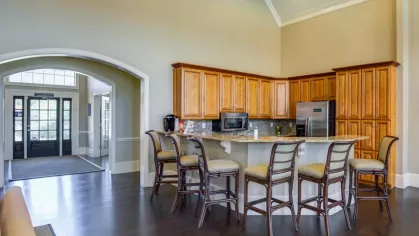 The community clubhouse kitchen, featuring wood cabinets, stainless steel appliances, and a granite breakfast bar. Barstools provide seating for residents.