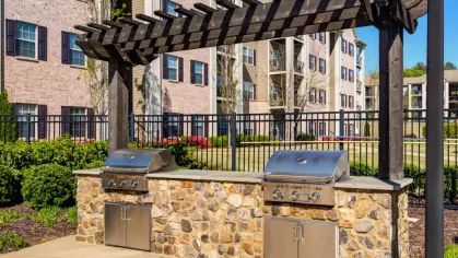 The outdoor grilling station features two stainless steel grills built into a stone counter. A dark wood pergola provides shade for the community amenity.