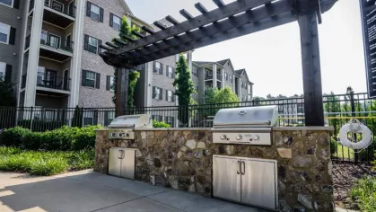 A wide view of the outdoor community grilling station, with two stainless steel grills in a stone base under a dark wood pergola.