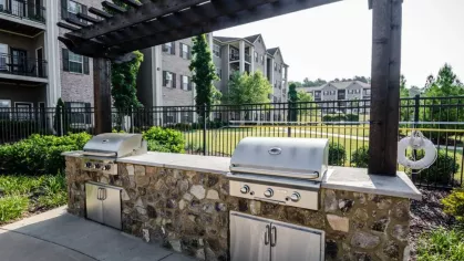 A side view of the outdoor grilling station under a pergola. The amenity is next to a fenced-in community green space and apartment buildings.