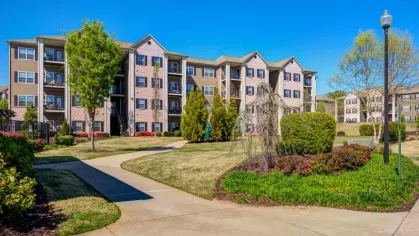 A paved walkway curves through a landscaped community green space. A large apartment building with private balconies is visible in the background on a sunny day.