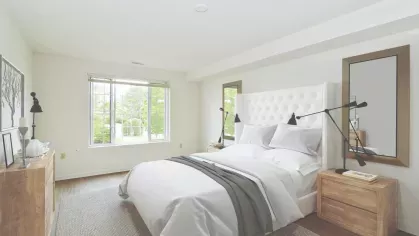 Serene bedroom with white upholstered headboard, matching nightstands, and window views of lush greenery outside.
