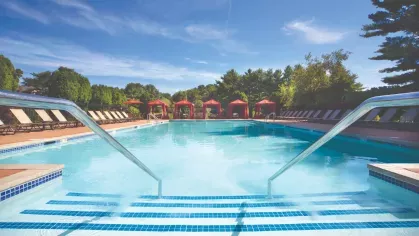 Resort-style pool with entry steps, red cabanas, and lounge seating framed by trees and blue skies.