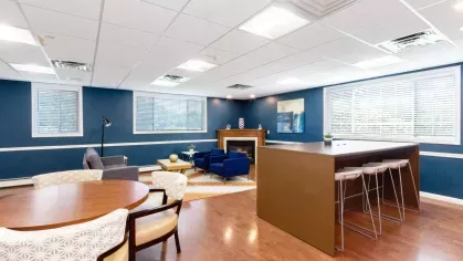 Clubhouse seating area with fireplace, blue accent chairs, and large windows bringing in natural light.