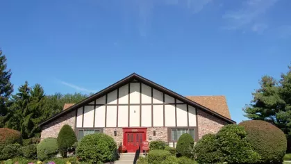 Leasing office entrance framed by colorful flowerbeds and trimmed bushes against a bright blue sky.