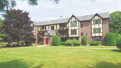 Apartment community with Tudor-inspired architecture, red entry doors, and lush landscaping framed by tall evergreens.
