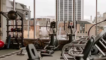 A modern apartment fitness center with cardio machines, weight equipment, and large windows overlooking the downtown city skyline.
