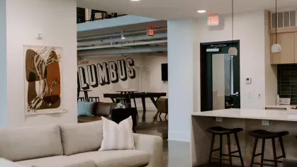 Modern resident lounge with cozy seating, a kitchen island, barstools, and a view into the community game room with a large “Columbus” mural.