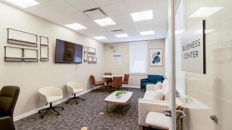 Modern business center with a cream sofa, assorted armchairs, a small meeting table, and a wall-mounted TV.
