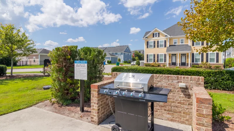Stainless steel gas grill in a brick enclosure, outdoors on a sunny day with green grass and a yellow house nearby.