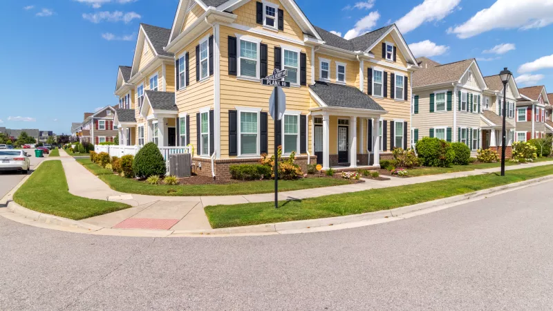 Row of colorful townhouses, including a prominent yellow one with black shutters, on a sunny street corner with green lawns.