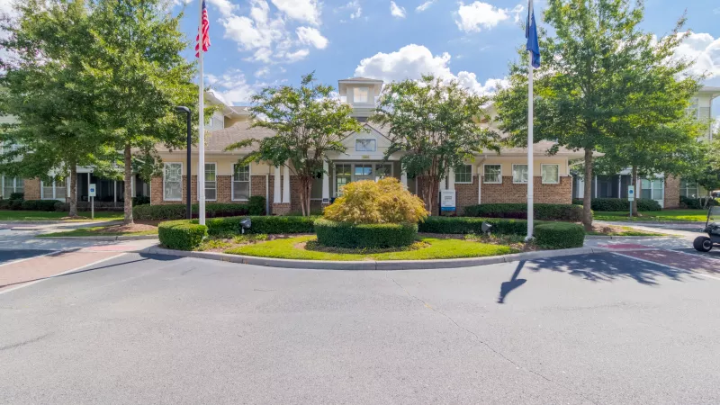 Community building entrance with a circular drive, two flagpoles, manicured shrubs, and green trees on a sunny day.