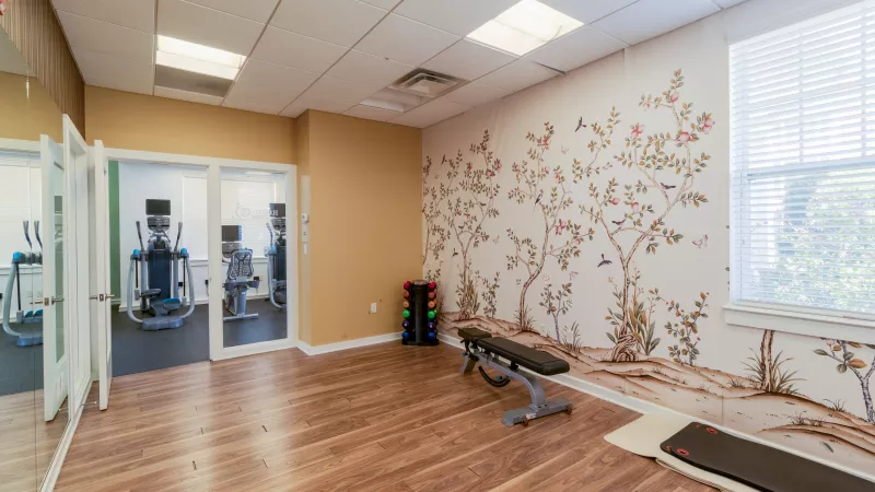 Fitness room with a nature mural, wooden floor, a weight bench, dumbbells, and cardio equipment in an adjoining space.