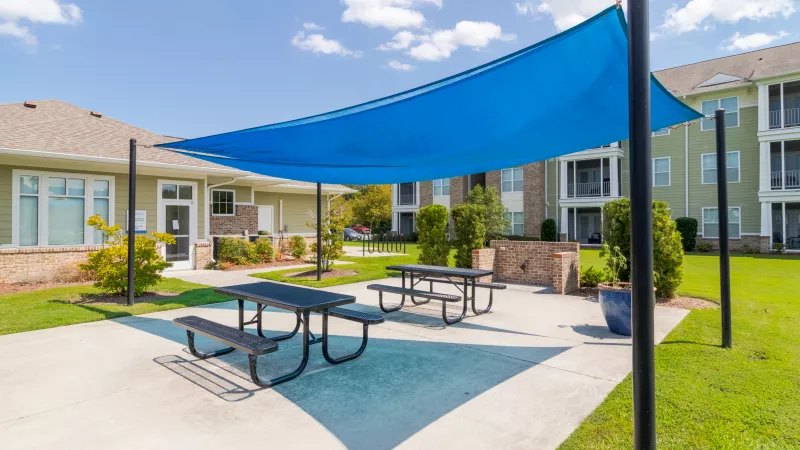 Sunny apartment complex patio with a large blue shade sail, two picnic tables, and green lawn.