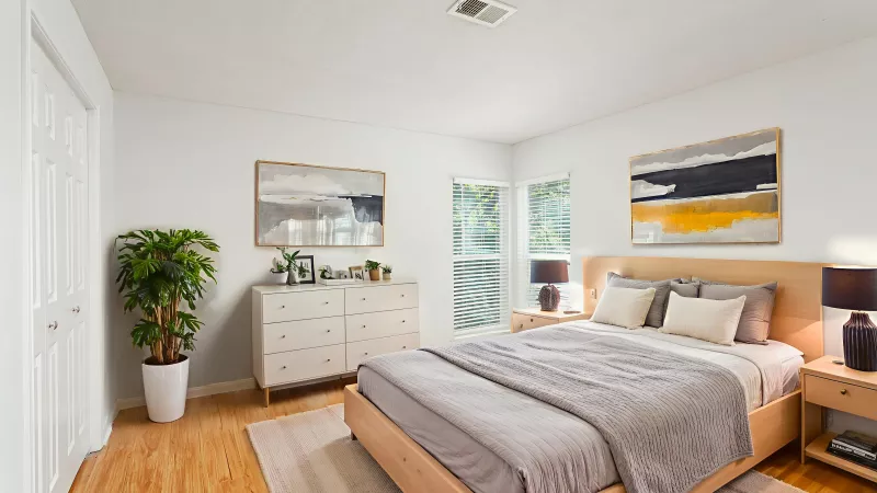 Bright bedroom with light wood bed, grey bedding, white dresser, tall green plant, and modern abstract art.