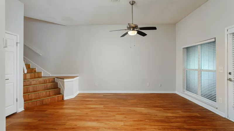 Empty living room with wood floors, light gray walls, brown-stepped staircase, ceiling fan, large window, and white door.