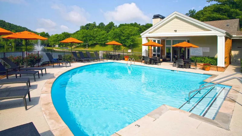 Outdoor swimming pool with blue water, orange umbrellas, and lounge chairs. A building with a patio, trees, and a pond are visible.