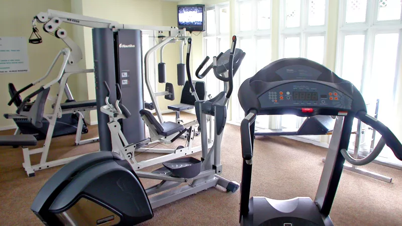 Small, well-lit gym with treadmill, elliptical, and weight machines. Large windows line the wall, and a TV is mounted above.