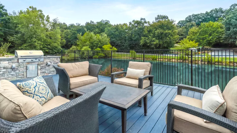 Patio with modern outdoor furniture on a blue deck, a built-in grill, and a pond surrounded by lush trees.