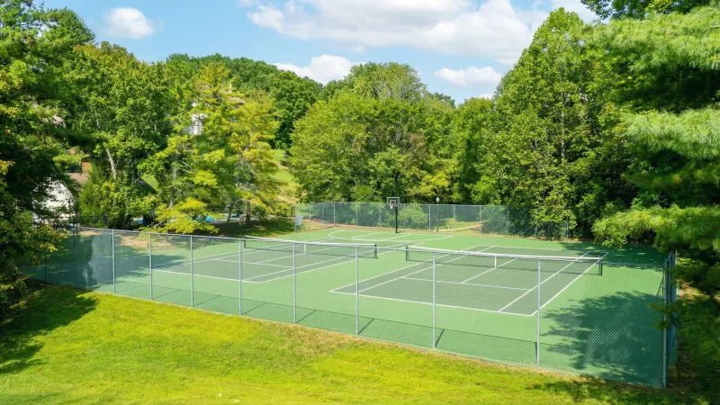 A pair of empty green tennis courts with fences, surrounded by vibrant green trees under a clear blue sky.
