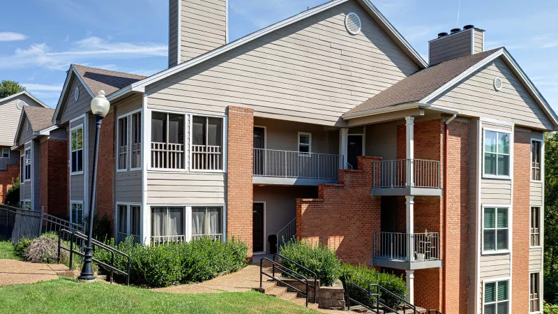 Multi-story apartment building with tan siding, brick, balconies, and green landscaping under a clear blue sky.