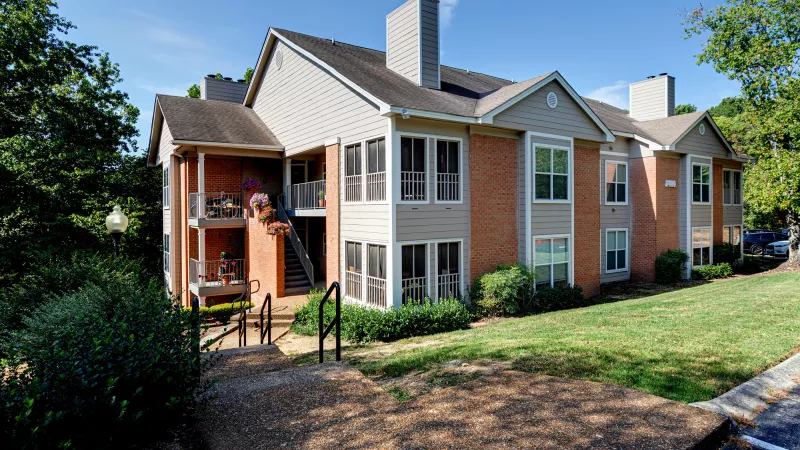 Three-story apartment building with light siding and brick, balconies, and a green lawn under a blue sky.