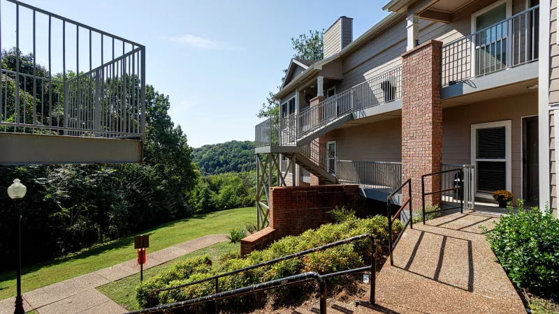 Light-colored residential building with balconies, ramps, and stairs, set against a backdrop of green, forested hills on a sunny day.