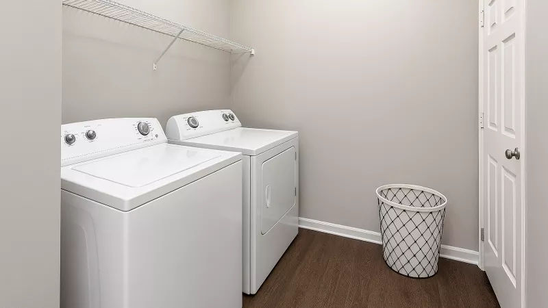 Modern laundry room with light beige walls, dark wood floor, two white top-load washers, wire shelf, and mesh hamper.