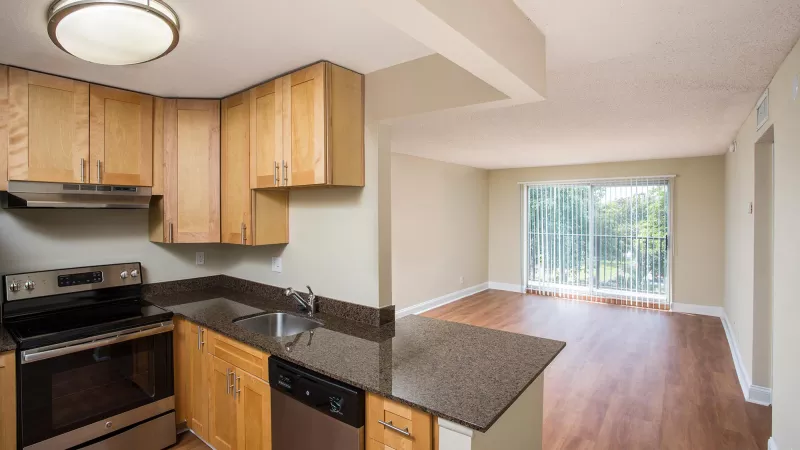 Kitchen with light wood cabinets, dark counters, stainless appliances, opening to a living room with wood floor and balcony.