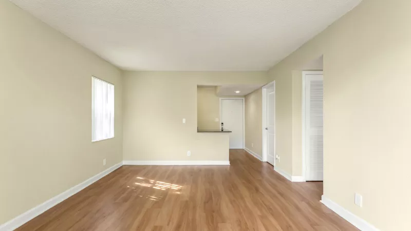 Spacious, empty living room with light walls, wood floor, window, and kitchen pass-through.