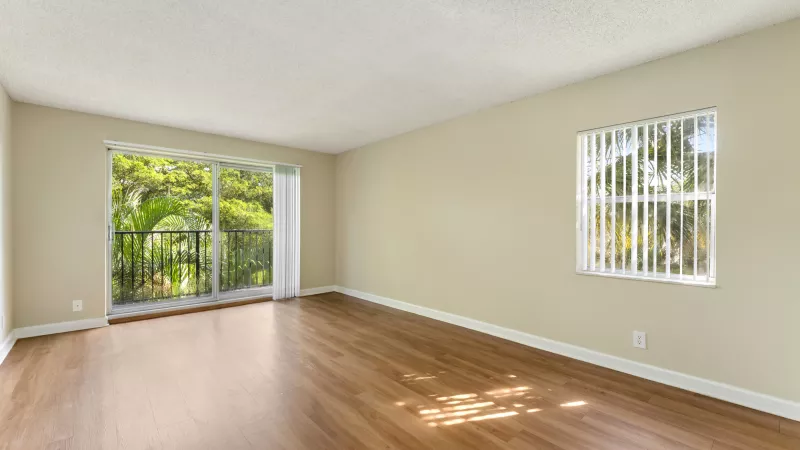Empty room with light beige walls, polished wood floor, a sliding glass door to a balcony, and a window with blinds.