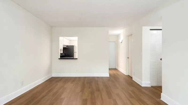 Empty living room with light walls, wood floor, kitchen pass-through on left, and hallway on right.