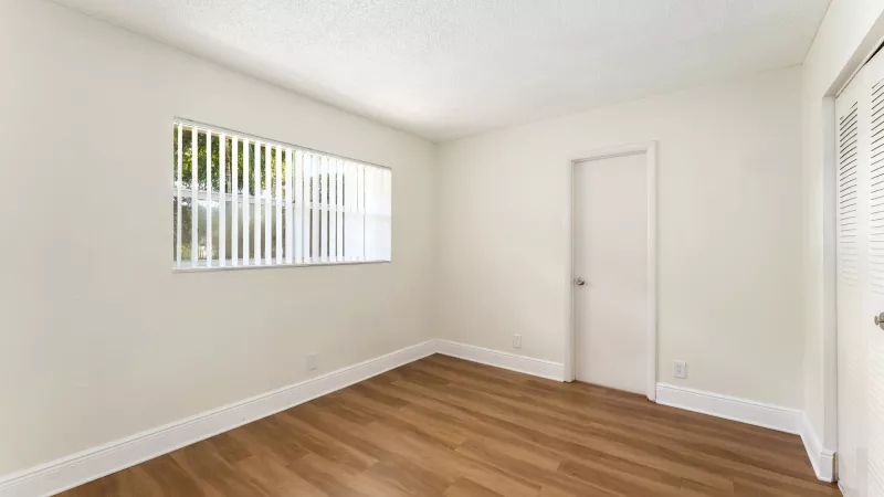 Empty room with light walls, wood-look floor, window with vertical blinds, and a closed white door.