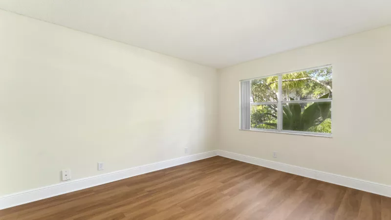 Empty room with light cream walls, wood laminate flooring, and a window overlooking lush green trees.