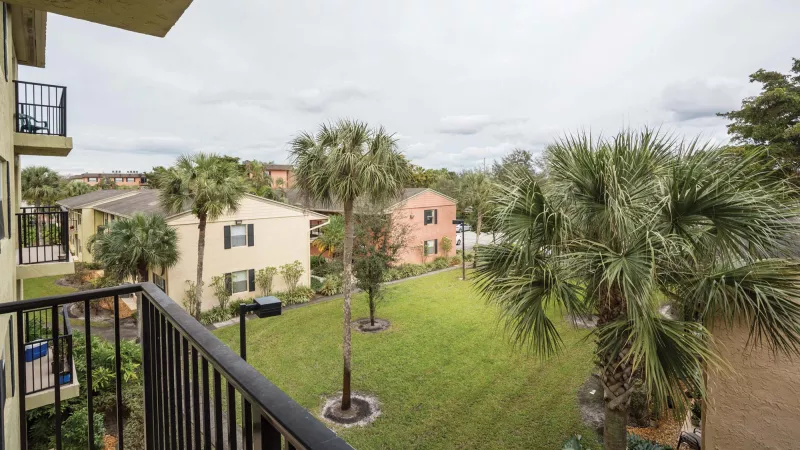 Balcony view of a green courtyard with palm trees and colorful apartment buildings under an overcast sky.