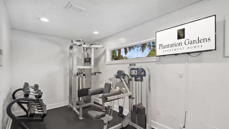 Bright gym with white walls, dark floor, various weight machines, dumbbell rack, and TV displaying "Plantation Gardens" logo.