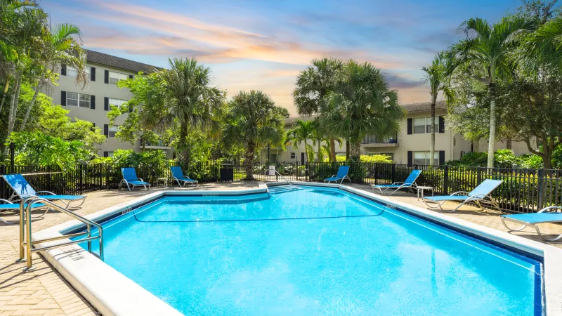Bright blue swimming pool surrounded by blue lounge chairs, palm trees, and apartment buildings under a partly cloudy sky.