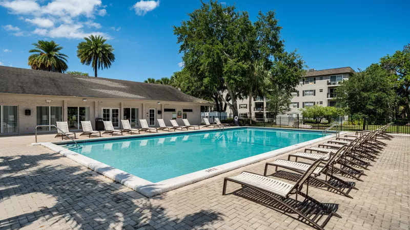 Outdoor swimming pool with blue water on a sunny day, surrounded by lounge chairs, buildings, and green trees.