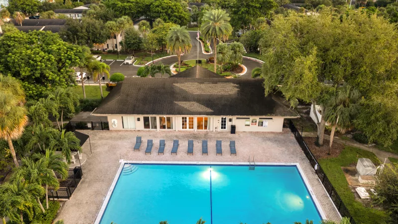 Aerial photo of a vibrant blue swimming pool, clubhouse, and lounge chairs among lush palm trees.