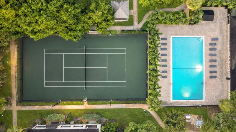 Photograph: Overhead view of a green tennis court and a vibrant blue swimming pool with sun loungers, nestled in trees.