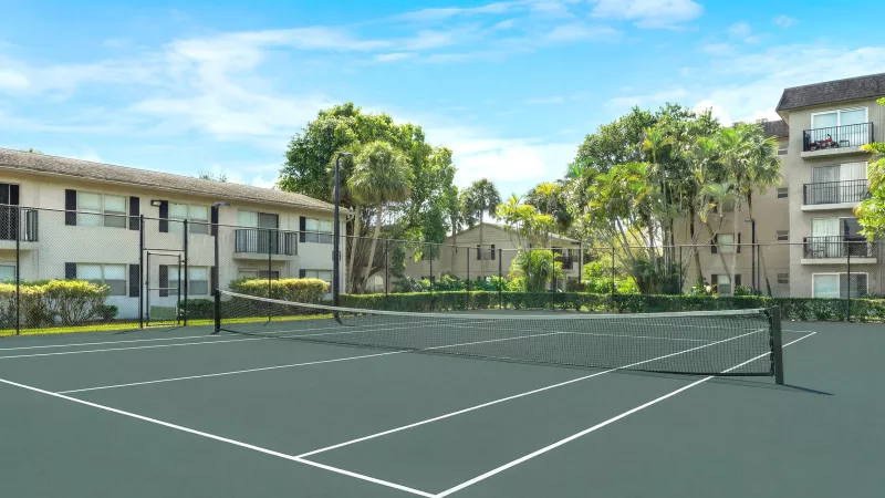Green tennis court with net, apartment buildings, and palm trees under a bright blue sky.