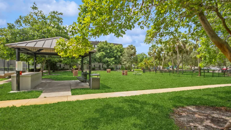 Lush green park featuring a covered gazebo with a grill station, a paved path, and abundant trees under a blue sky.