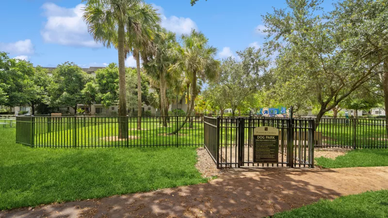 Photograph: Fenced dog park with lush green grass, palm trees, a dirt path, and an informational sign.
