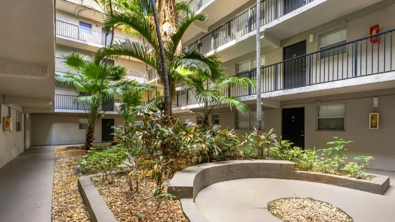 Photograph of a tropical apartment complex courtyard with palm trees, lush plants, a curved concrete bench, and gravel.